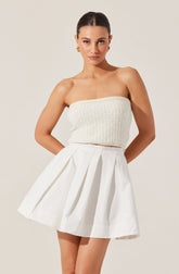 A woman is wearing a strapless, knitted, cream-colored top paired with a high-waisted, white pleated skirt. She has her hair styled in a sleek, pulled-back look and is accessorized with gold earrings.